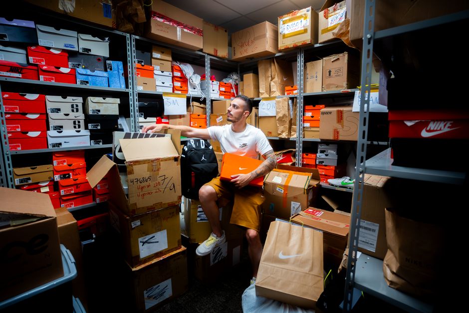 A man organizing various shoe boxes in a cluttered store room filled with shelves and cartons.